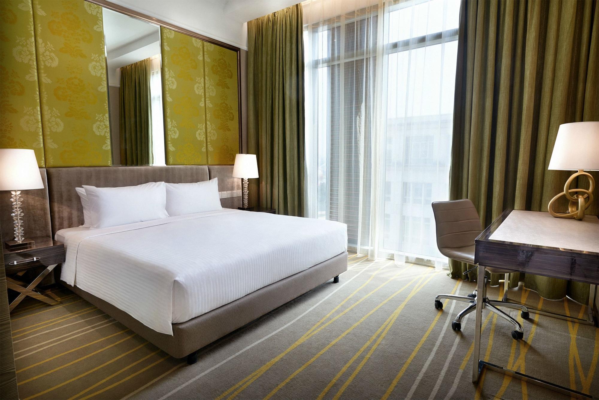 Dorsett Putrajaya Hotel Exterior photo The photo shows a modern hotel room. It features a large bed with white bedding positioned against a wall. The room has tall windows covered with sheer curtains, letting in natural light. There are two bedside lamps on either side of the bed. A desk 