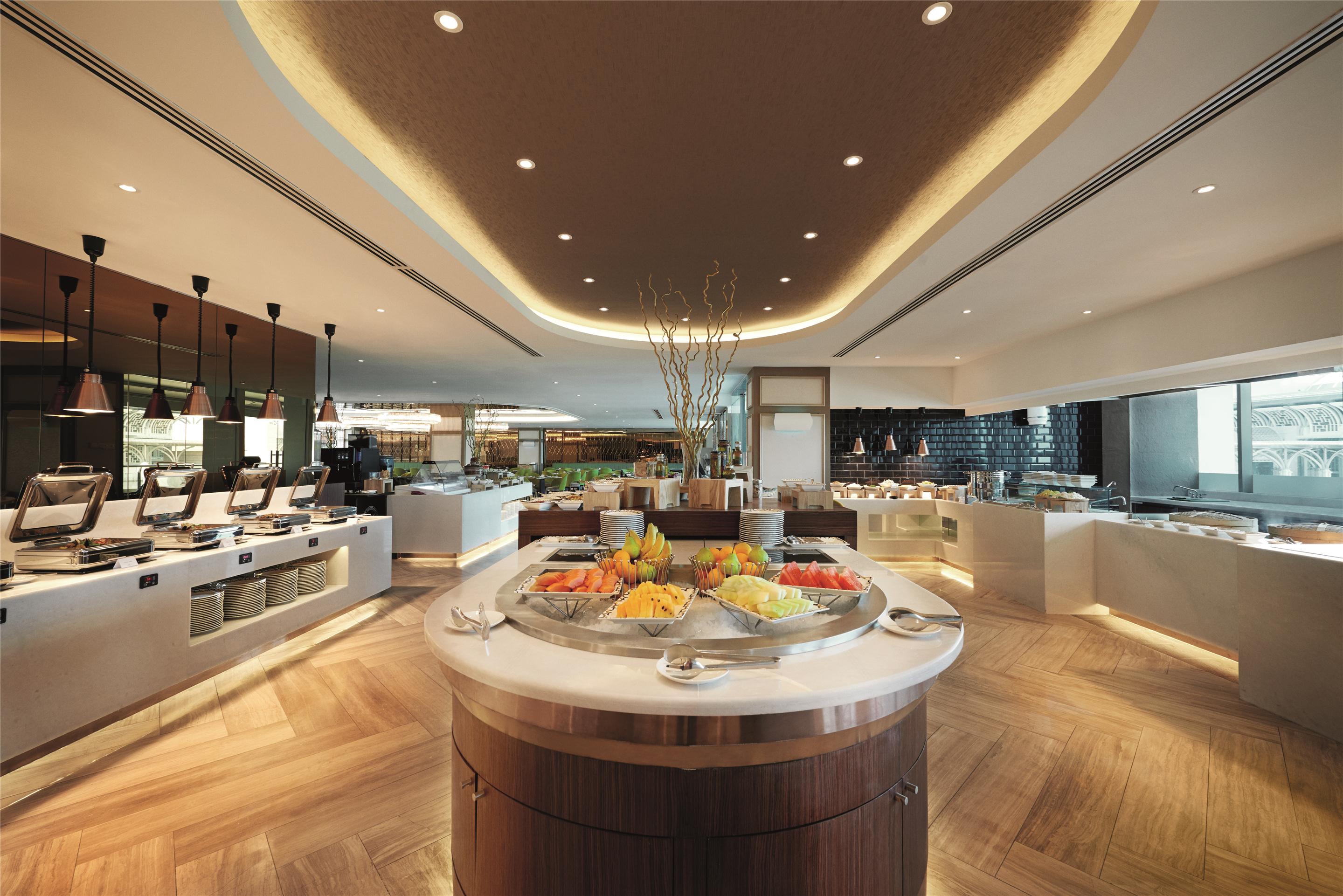 Dorsett Putrajaya Hotel Exterior photo The photo shows a modern buffet restaurant interior. In the center, there's a circular display with a variety of fresh fruits arranged attractively. Surrounding this are several serving stations and food displays that feature a clean, contemporary de
