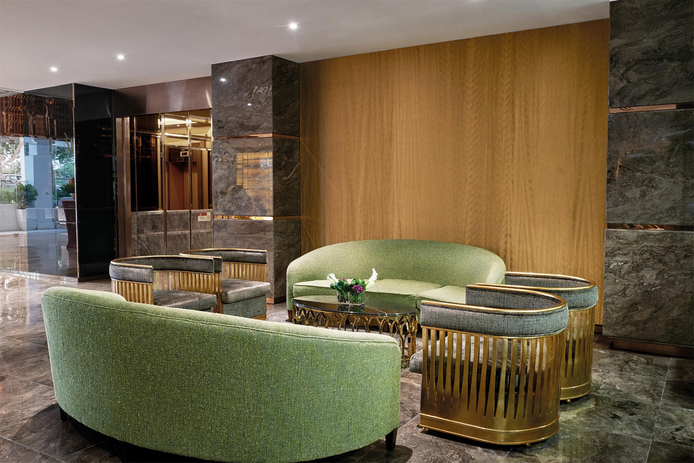 Dorsett Putrajaya Hotel Exterior photo The photo shows a stylish lounge area with modern furniture. There is a large, curved green sofa accompanied by matching green accent chairs. The seating is arranged around a unique gold and glass coffee table. The backdrop features a wooden wall wit