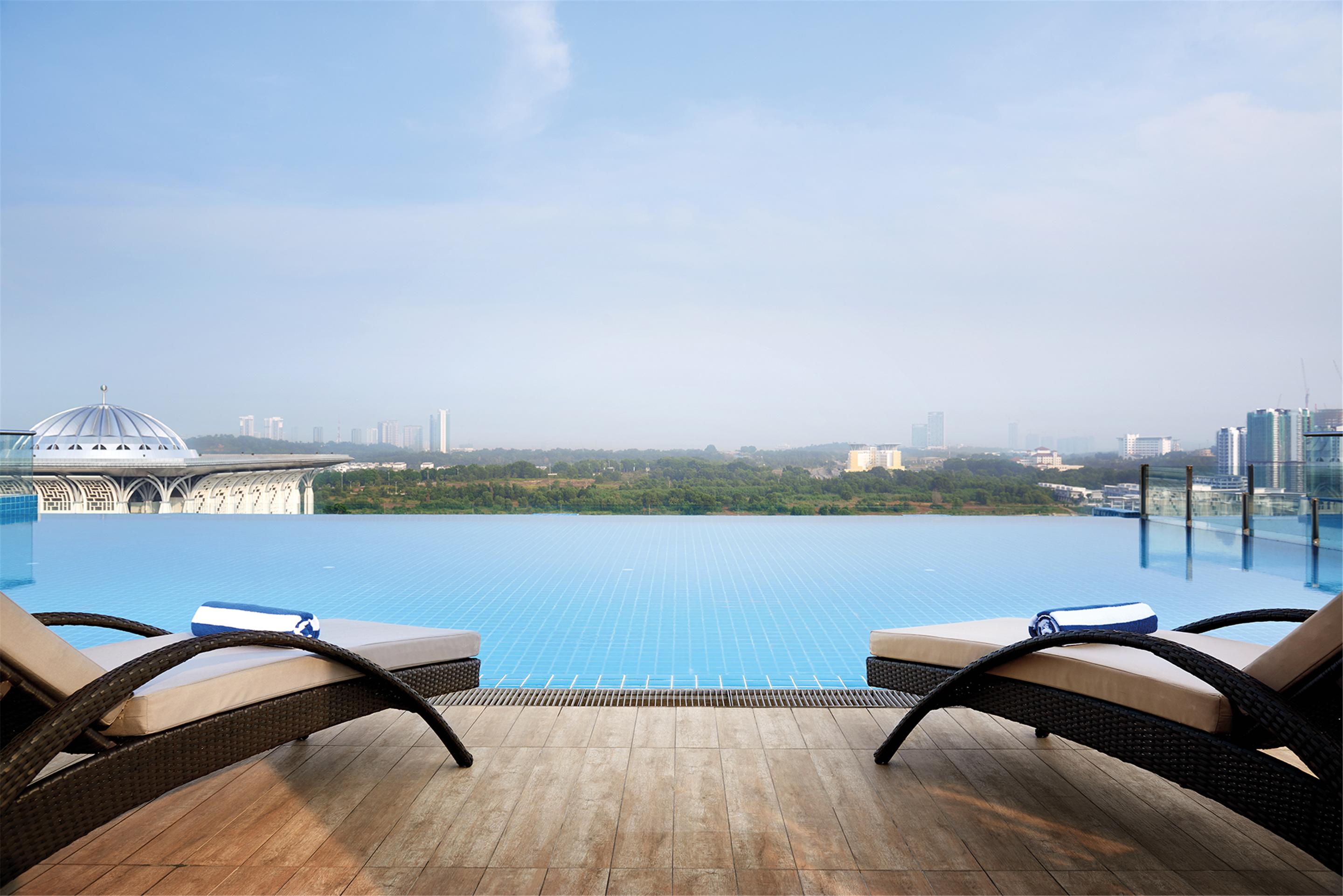 Dorsett Putrajaya Hotel Exterior photo The photo depicts a luxurious outdoor space featuring an infinity pool that extends toward the horizon, blending seamlessly with the sky. In the foreground, there are two sun loungers with rolled towels on each, positioned on a wooden deck. The backd