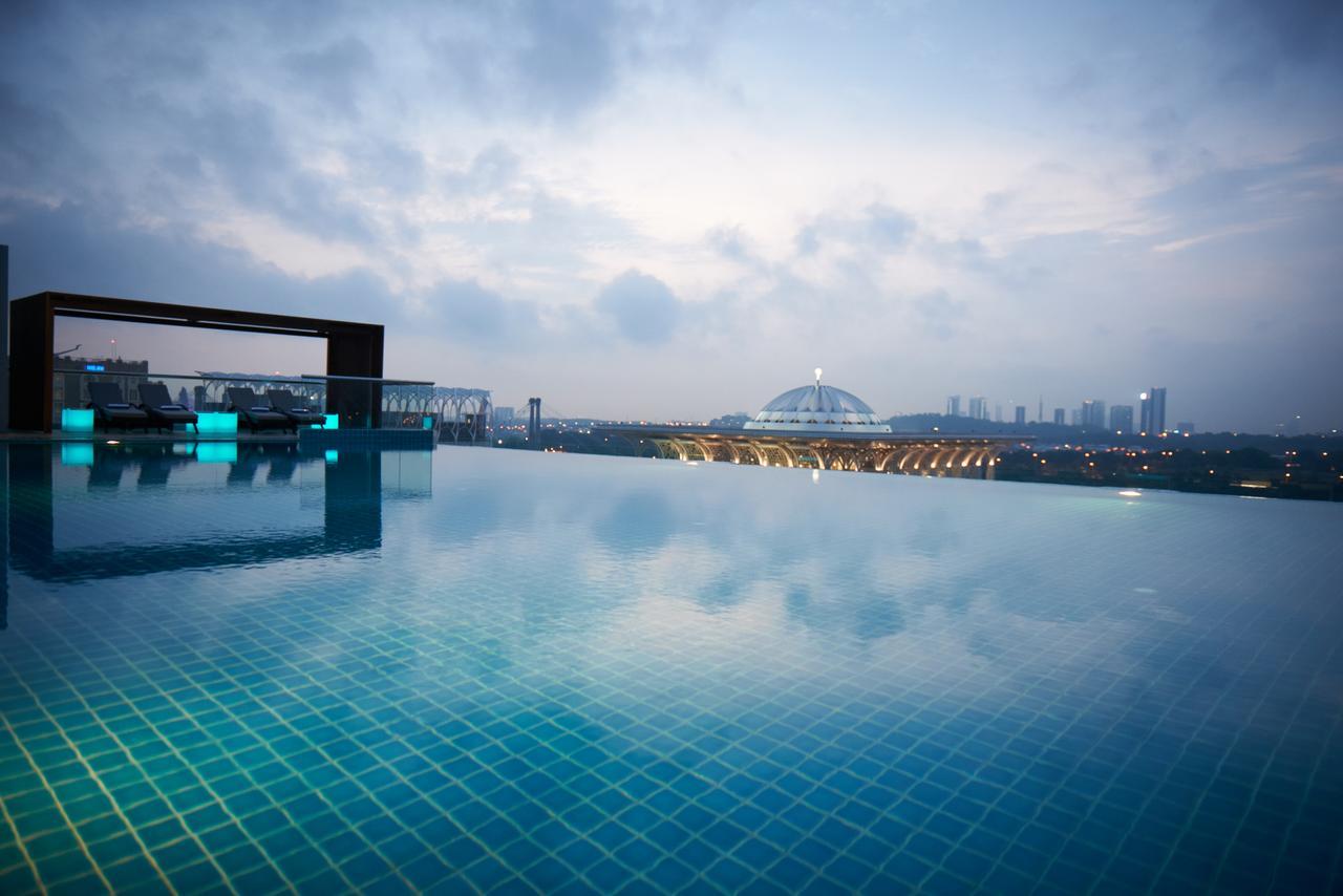 Dorsett Putrajaya Hotel Exterior photo The photo shows a serene infinity pool with a calm, reflective surface, blending seamlessly with the sky. In the background, there's a view of a cityscape with buildings and a large dome structure, possibly a stadium or an arena. The atmosphere appea