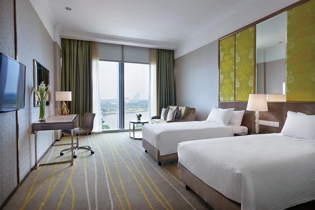 Dorsett Putrajaya Hotel Room photo The photo shows a modern hotel room featuring two single beds with white linens. There is a stylish desk and chair positioned beside a large window that allows natural light to fill the space. The window is adorned with long, light green curtains. A 