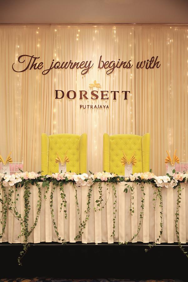 Dorsett Putrajaya Hotel Exterior photo The photo features a decorative setup, likely for an event. In the center, there are two prominent yellow chairs with tufted upholstery, positioned against a backdrop of soft draping. Above the chairs, the name "DORSETT PUTRAJAYA" is displayed, indic