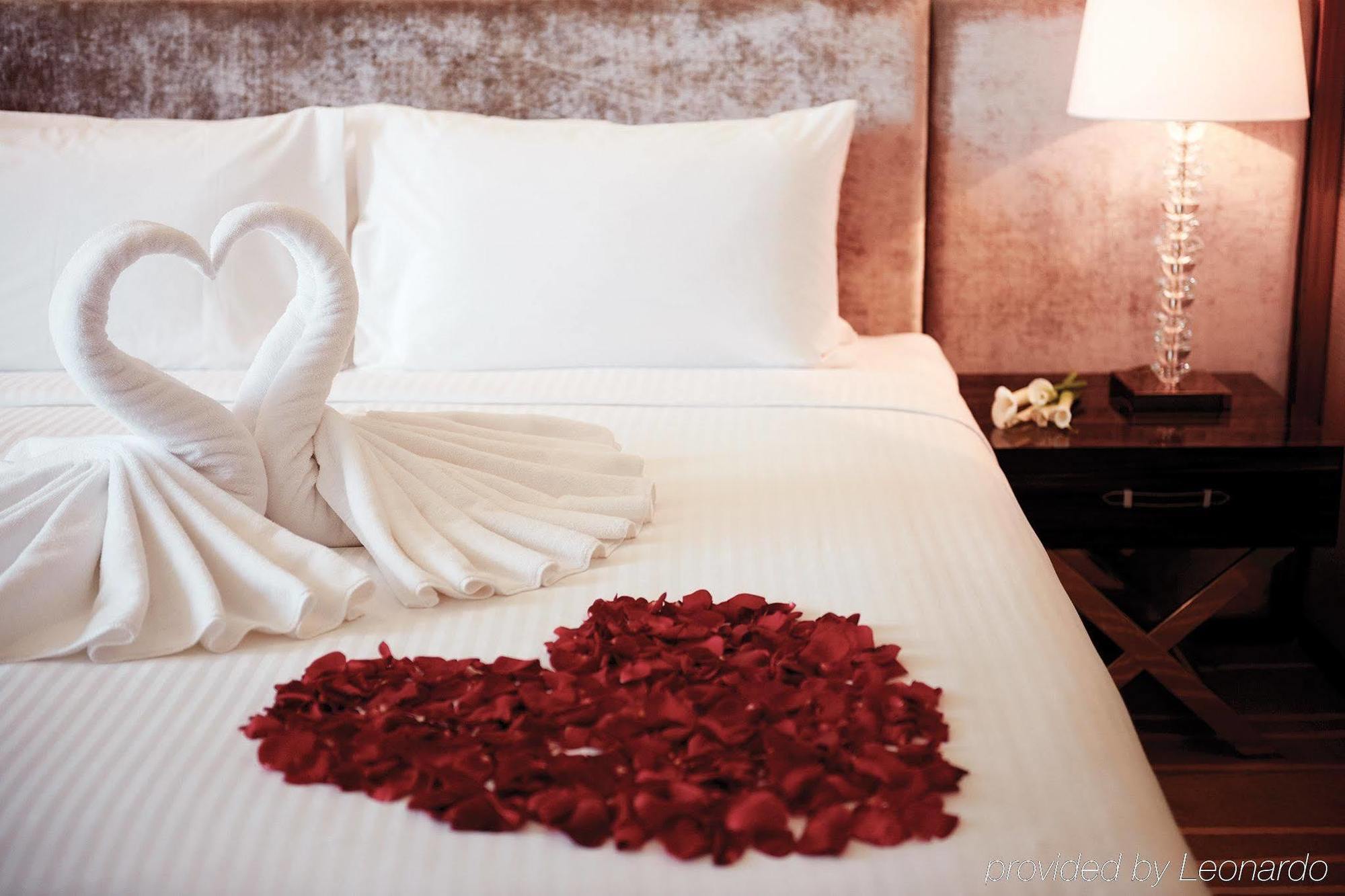 Dorsett Putrajaya Hotel Exterior photo The photo displays a romantic hotel bed setting. On the neatly arranged white sheets, two swan-shaped towel sculptures are positioned, resembling a heart. Surrounding the towels, there is a heart shape made of red rose petals. To the side, there is a