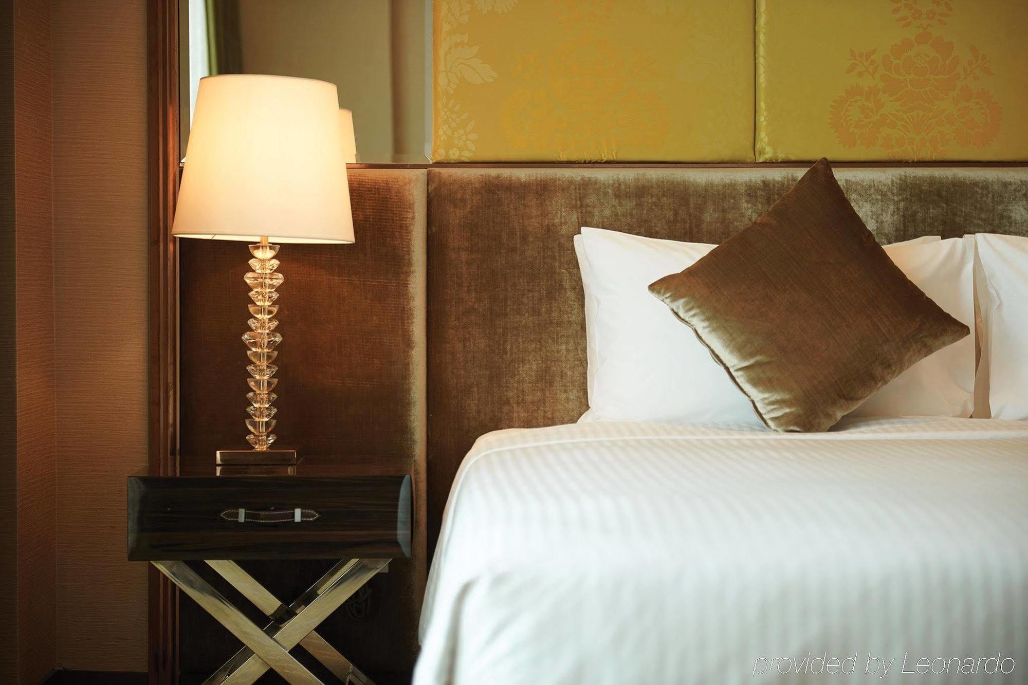 Dorsett Putrajaya Hotel Exterior photo The photo features a cozy bedroom setting. There is a bed with a neatly arranged white bedsheet and a decorative brown cushion placed against a soft, upholstered headboard. Beside the bed, there's a stylish bedside table with an elegant lamp that has