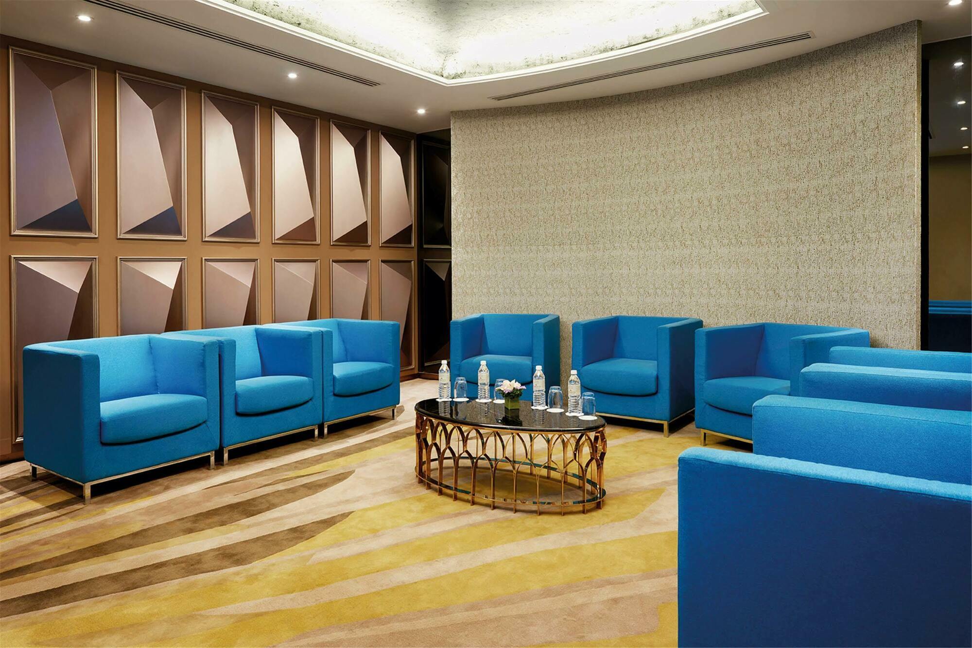 Dorsett Putrajaya Hotel Exterior photo The photo shows a modern meeting or lounge area. It features bright blue chairs arranged around a central table. The table has decorative elements and bottles of water on it. The room is designed with stylish walls that have a geometric pattern and a