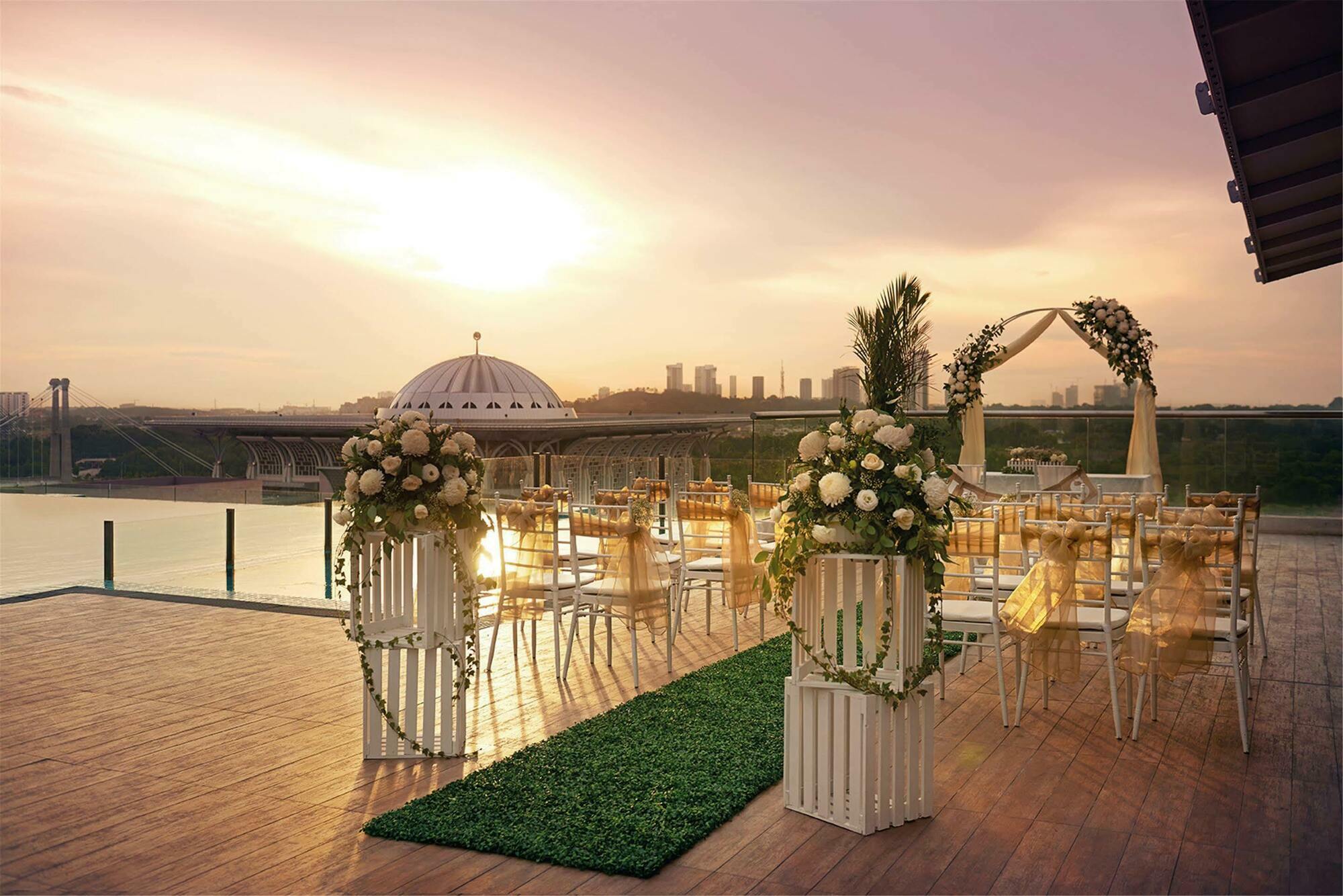 Dorsett Putrajaya Hotel Exterior photo The photo shows a beautifully designed outdoor wedding venue. In the foreground, there are elegant arrangements of flowers on white pedestals, creating a romantic atmosphere. There is a pathway lined with green grass leading to an arch adorned with f